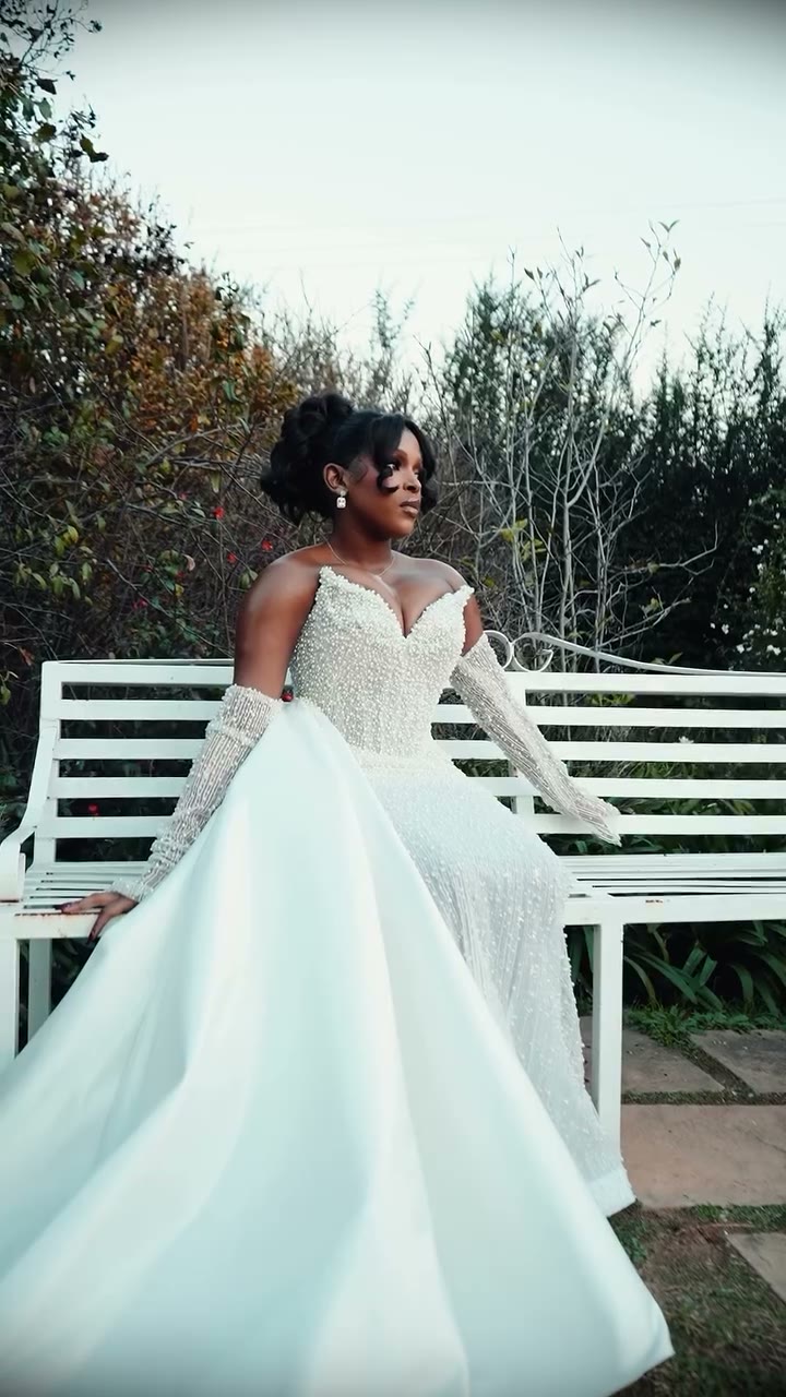 Bride seated on a white garden bench in a pearl-encrusted gown with a full satin overskirt and sheer pearl-beaded sleeves
